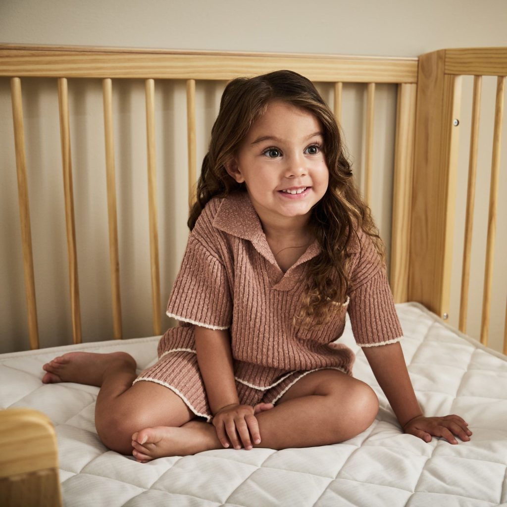 Toddler sitting on The Little Green Sheep Twist Natural Cot Bed Mattress in a cot bed