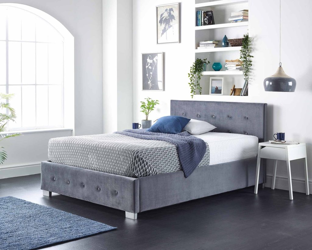 Aspire Side Opening Ottoman Storage Bed in grey velvet, three-quarter view in a bright bedroom