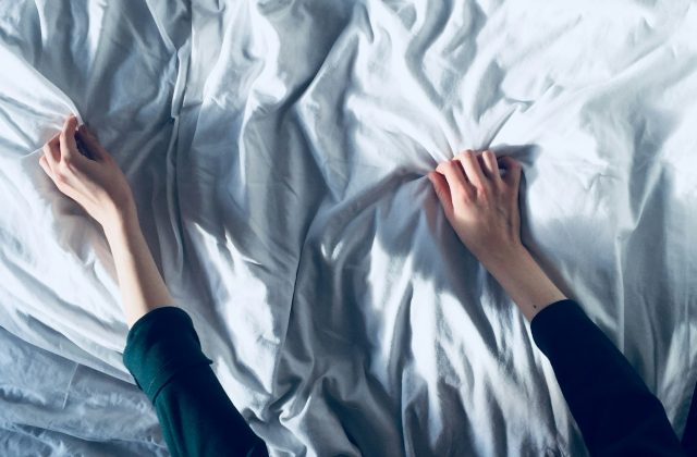 Two hands pulling back a crumpled white sheet to strip a bed before cleaning the mattress