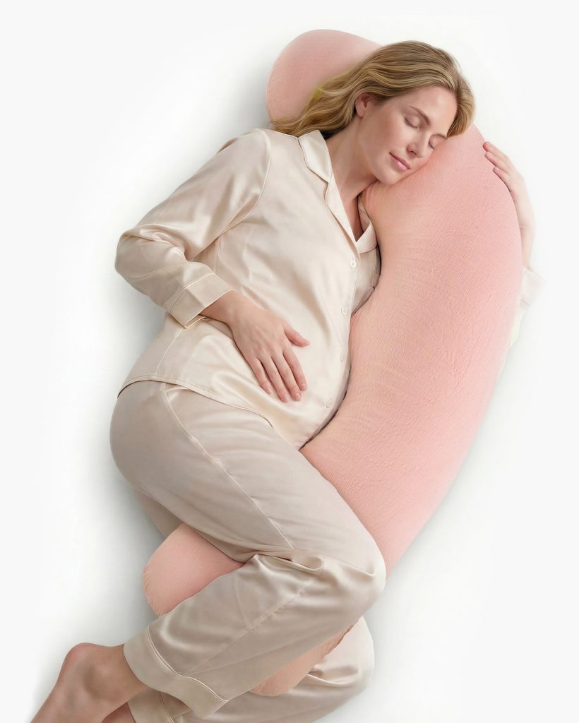 Pregnant woman in pyjamas resting on the Momcozy J-shaped pillow in the upgraded pink cotton cover
